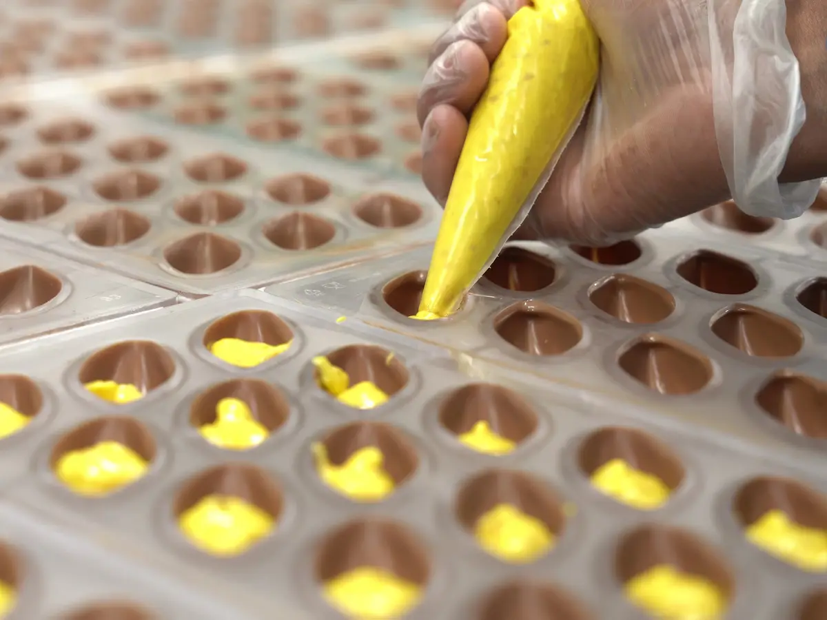 A gloved hand piping a golden filling into moulded chocolate shells